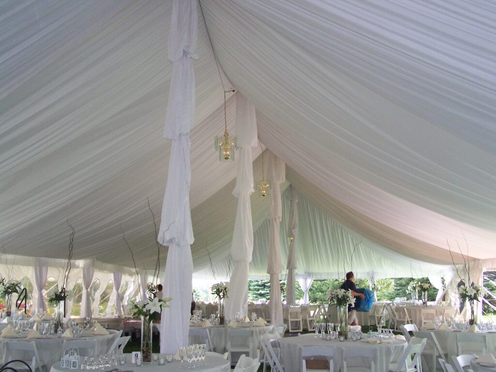 A large white tent set up for an event with round tables, white chairs, and decorative drapes and chandeliers.