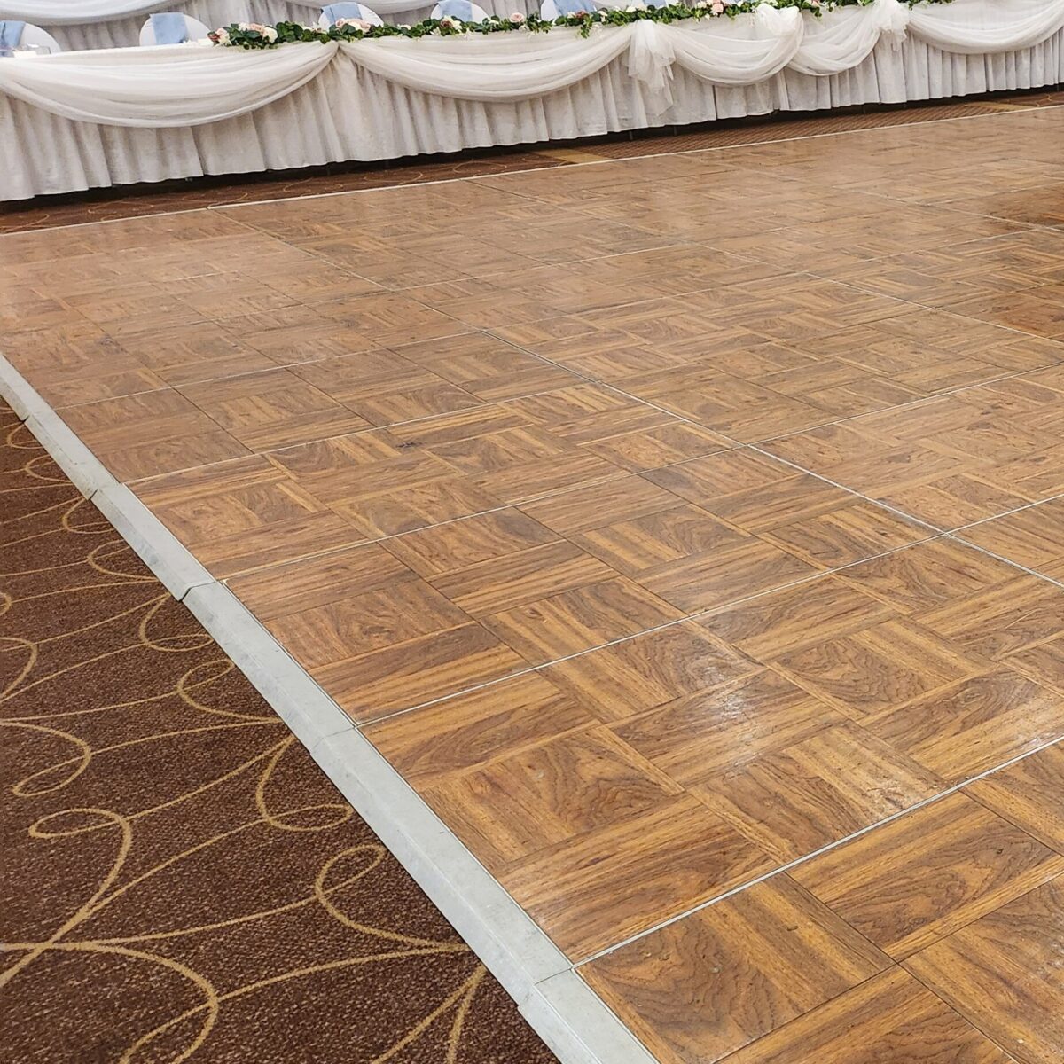 Empty dance floor with a wooden parquet design next to a carpeted area, in a decorated venue with draped tables in the background.