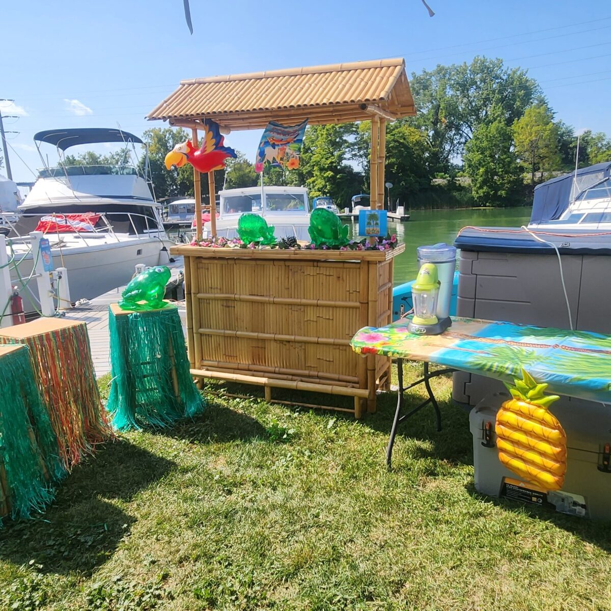 Tiki bar setup near a dock with boats, decorated with inflatable items, grass skirts, and a pineapple-themed cooler on grass.