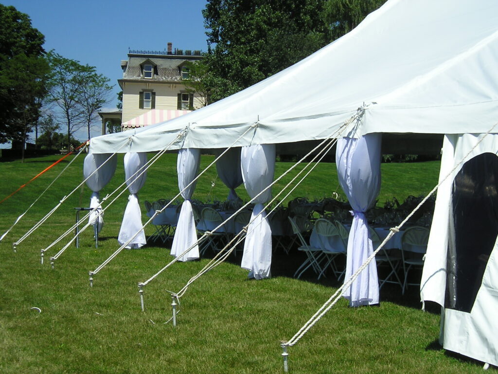 White outdoor event tent with round tables and chairs is set up on a grassy lawn near a house.