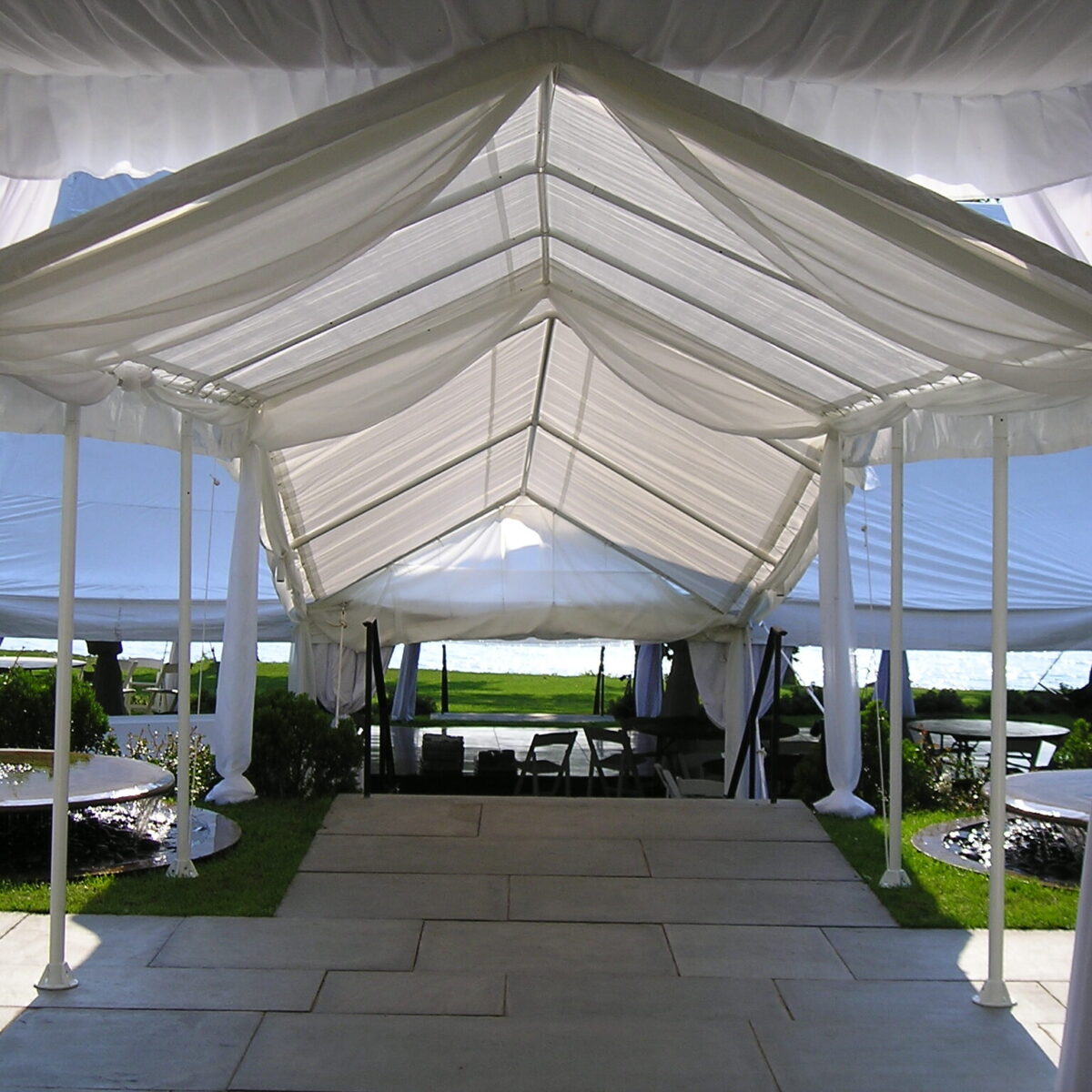 A white tent with a peaked roof covers a pathway, leading to an outdoor area with grass and a view of the water.