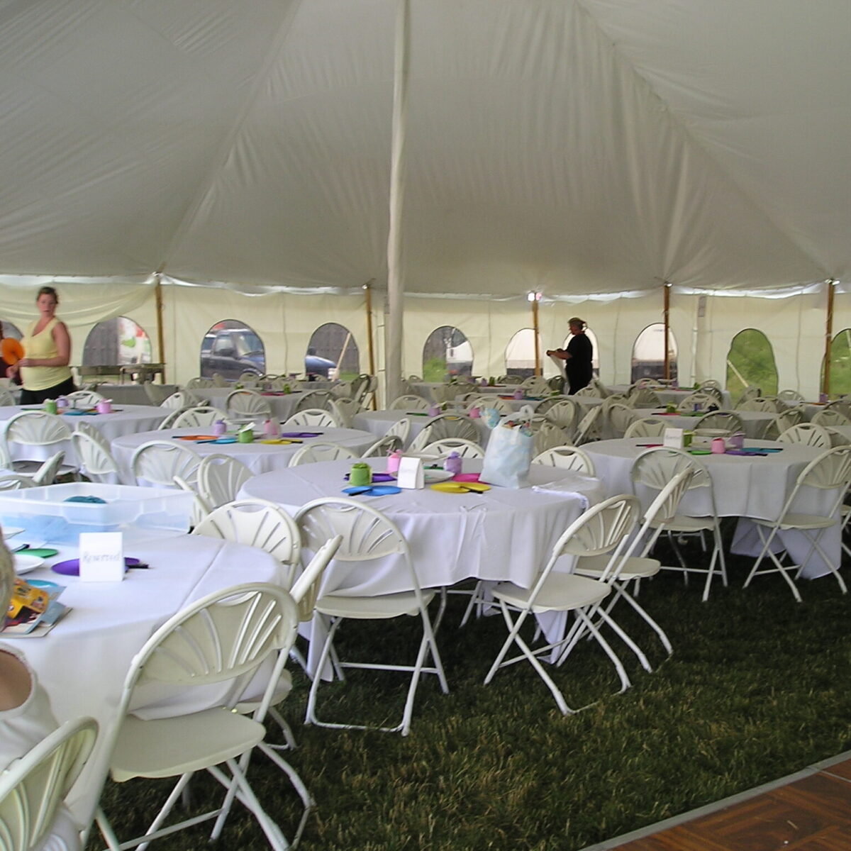 A large white tent with round tables covered in white cloths and surrounded by folding chairs. People are setting up the space.